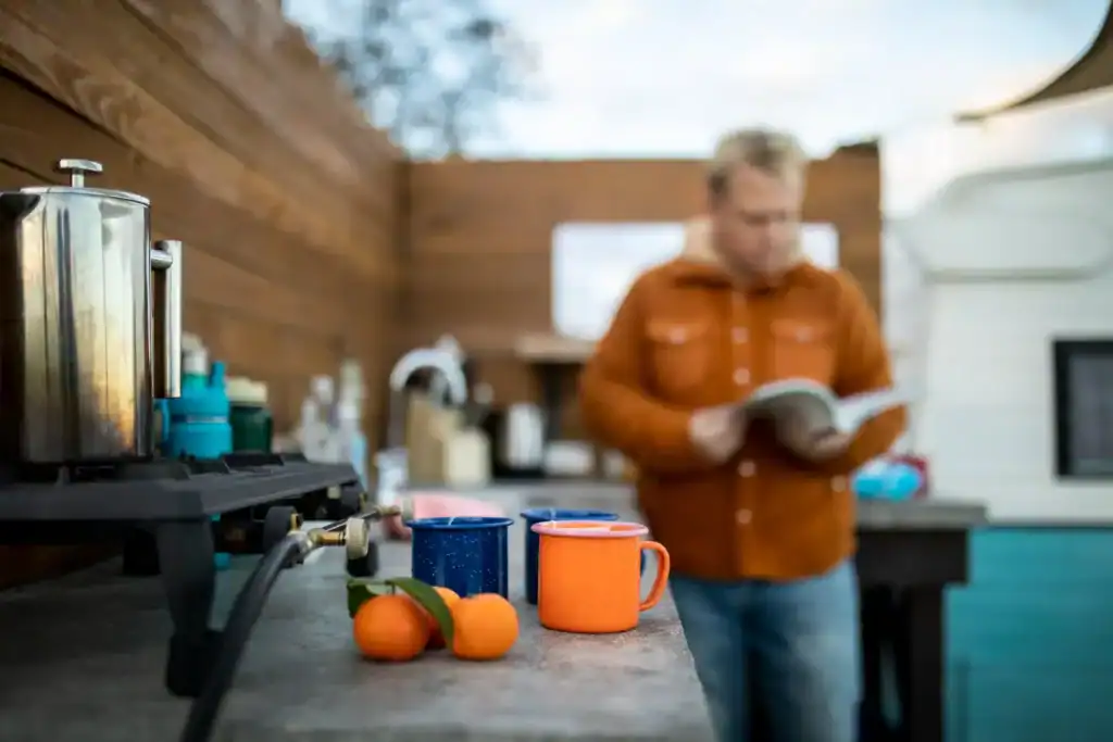 A person reading a book in a cozy outdoor kitchen with a wooden wall, coffee mugs, oranges, and a coffee maker creating a perfect relaxing space.