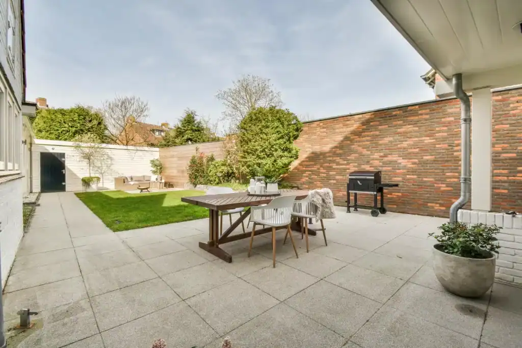 A spacious backyard patio featuring outdoor dining, lush greenery, a barbecue grill, and a brick wall, creating an inviting space for relaxation and entertaining.