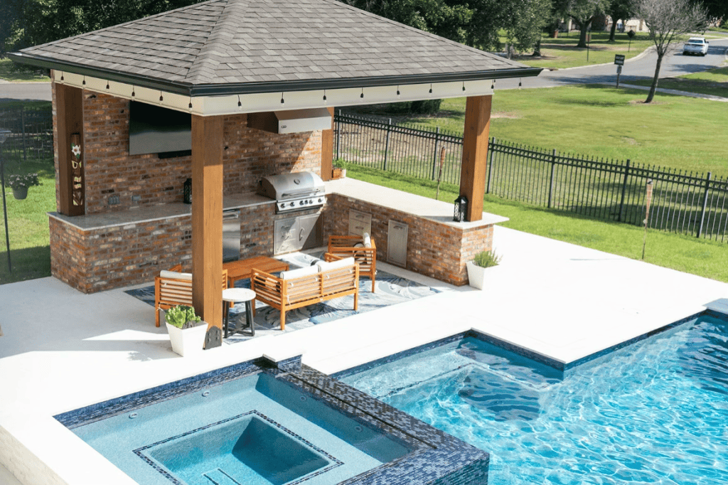 Elegant poolside outdoor kitchen with a built-in grill, seating area, and brick accents overlooking a sparkling swimming pool and hot tub in a lush yard.