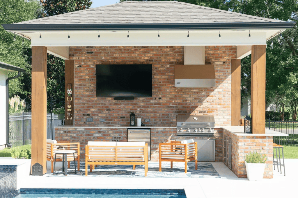Stylish outdoor kitchen and lounge area with brick wall and built-in grill at Duplessis Builders’ custom pool project.