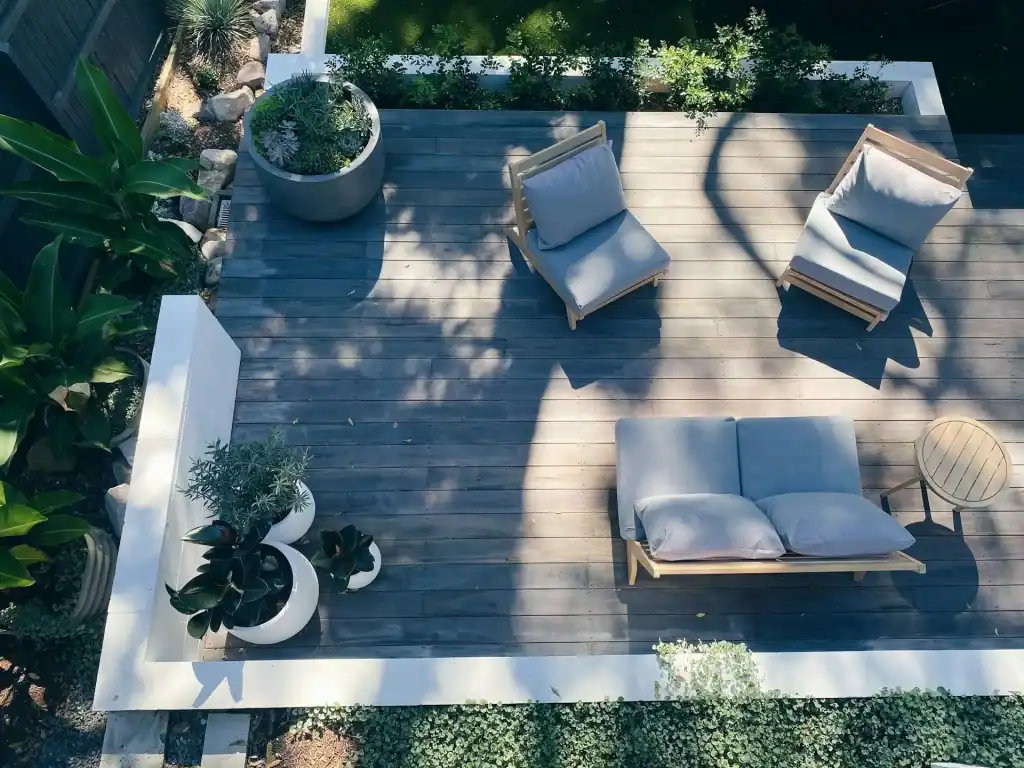 Wood deck with outdoor lounge furniture, potted plants, and lush greenery, creating a relaxing patio space next to a swimming pool.