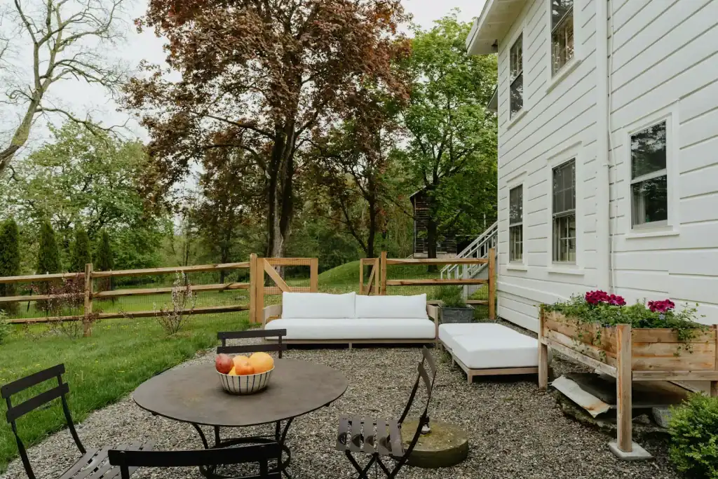 Cozy backyard seating area with a white outdoor sofa, black chairs, and a round table with fruit, surrounded by greenery and a wooden fence beside a white house.