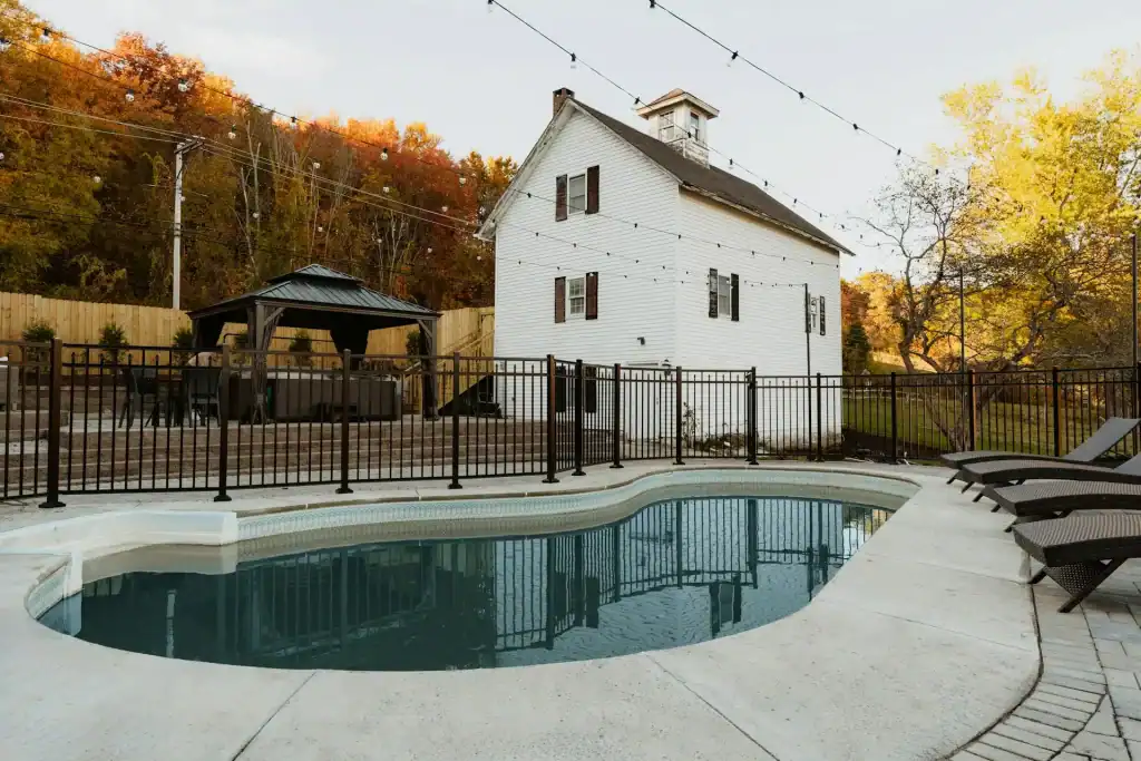 Pool with fenced perimeter, outdoor seating, and a white house in the background surrounded by fall foliage in a residential backyard.