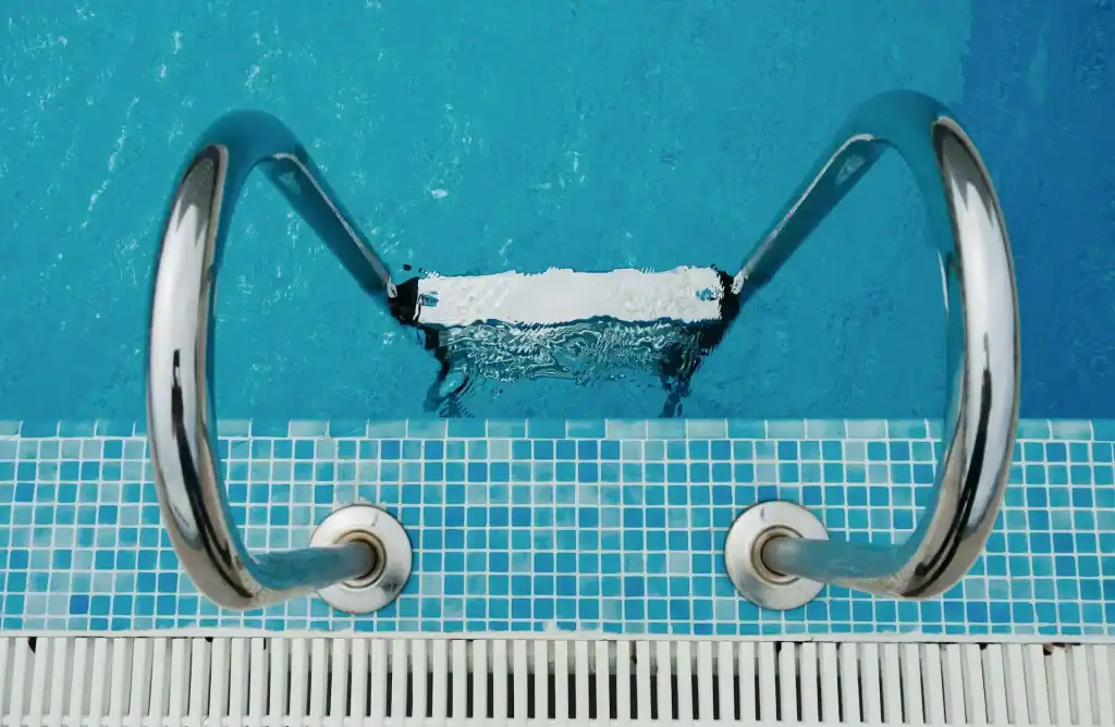 A stainless steel pool ladder descending into a clear blue swimming pool with small blue and white tile accents around the water's edge.