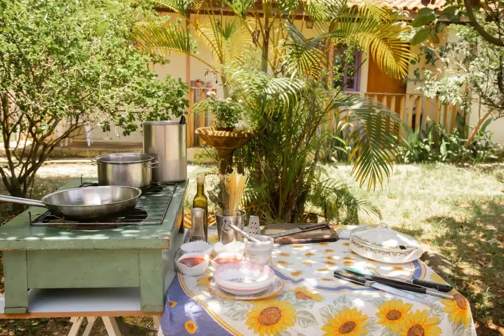 Fresh outdoor kitchen setup with cooking utensils, olive oil, and fresh pasta on a sunflower tablecloth in a lush garden. Perfect for summer poolside gatherings and outdoor cooking.