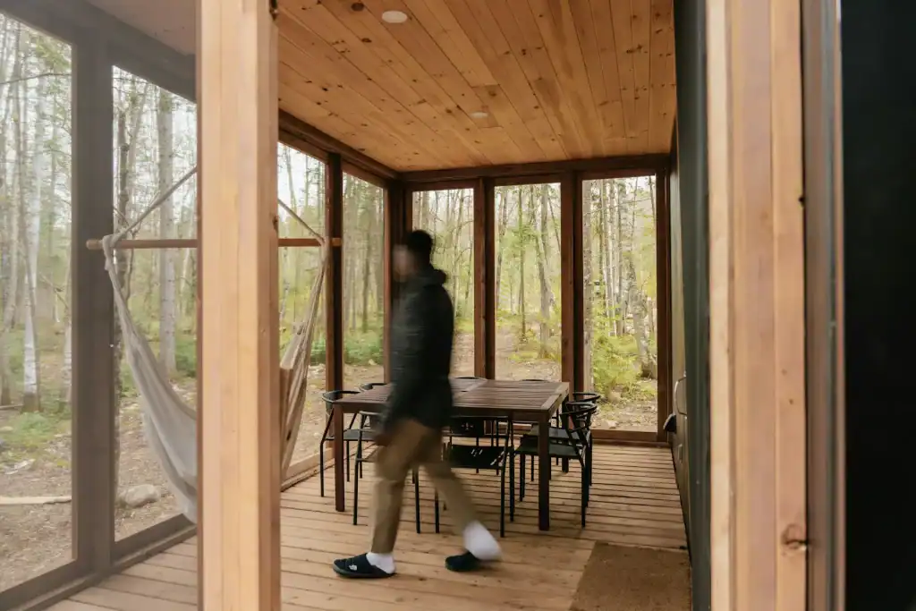 Modern screened-in porch with wooden accents, surrounded by lush forest, featuring a cozy hammock and a black dining table with chairs, perfect for outdoor relaxation and gatherings.