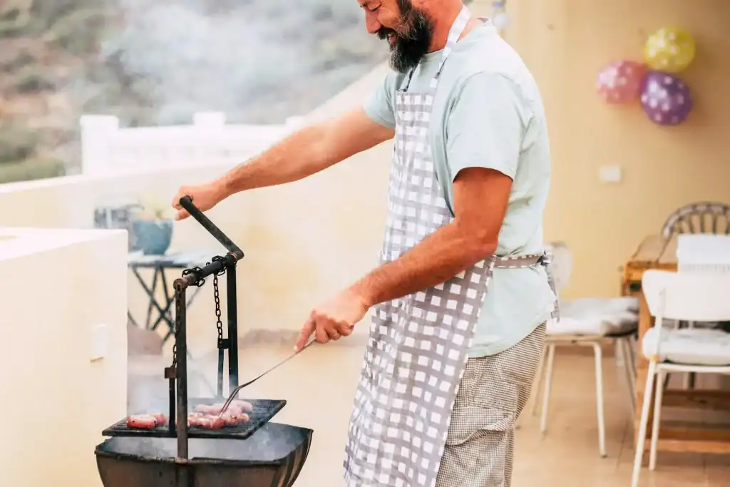 Grilling outdoors with a man cooking meat on a barbecue grill, wearing an apron, in a cozy indoor space with a warm, inviting atmosphere.