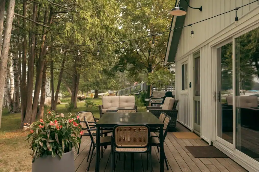 String lights hanging above an outdoor patio with a dining table, chairs, and a barbecue grill near a house surrounded by trees.