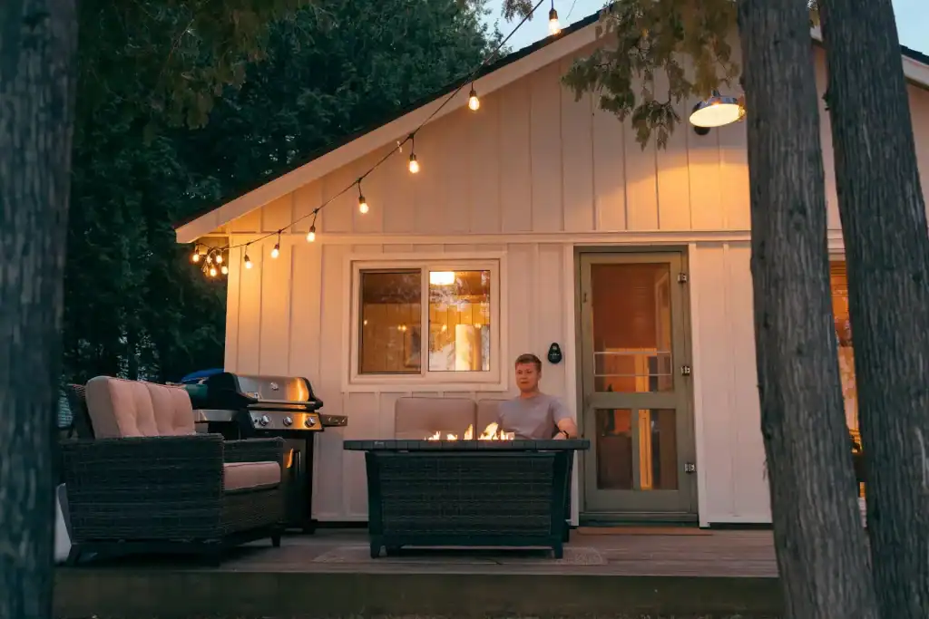 A cozy outdoor backyard seating area with a firepit, string lights, a built-in grill, and a modern small house surrounded by trees during the evening, perfect for relaxing and entertaining.