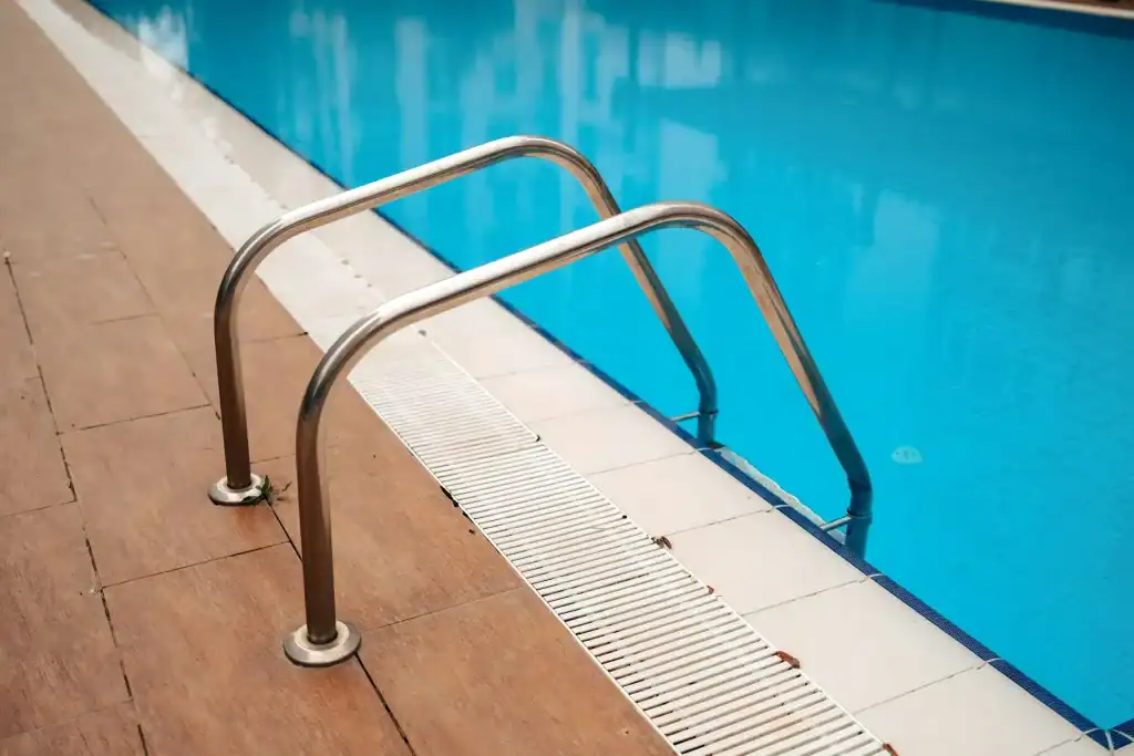 Elegant stainless steel pool ladder at the edge of a bright blue in-ground swimming pool with tile and drainage system.