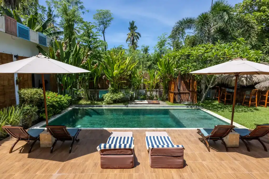 A beautiful tropical backyard pool area with lounge chairs, umbrellas, lush green plants, and tall trees under a clear blue sky, creating an ideal space for relaxation and outdoor enjoyment.