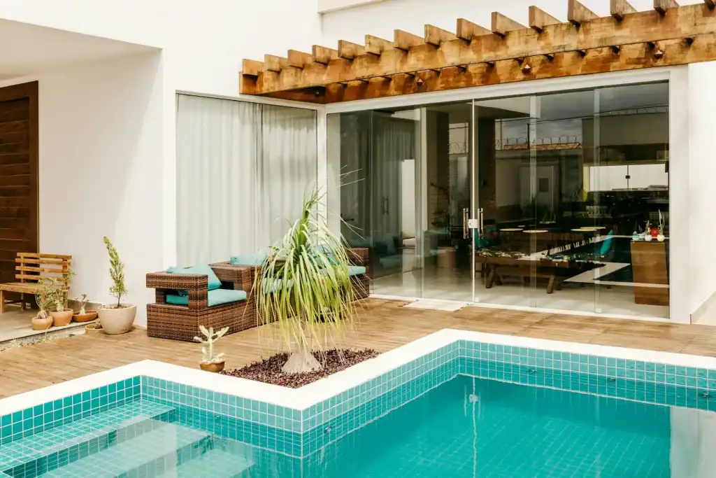 Luxurious backyard with a modern in-ground pool, wooden deck, potted plants, and a glass wall patio offering a stylish, relaxing outdoor living space.