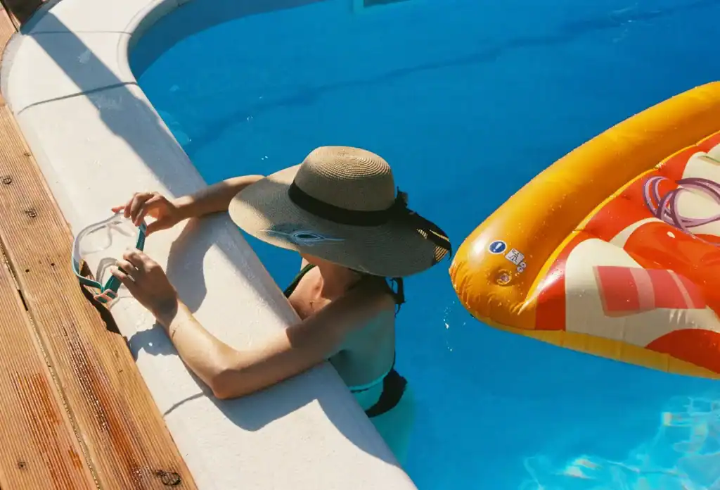 Relaxed woman with a wide-brimmed hat adjusting her snorkeling mask by a backyard swimming pool with a colorful float.