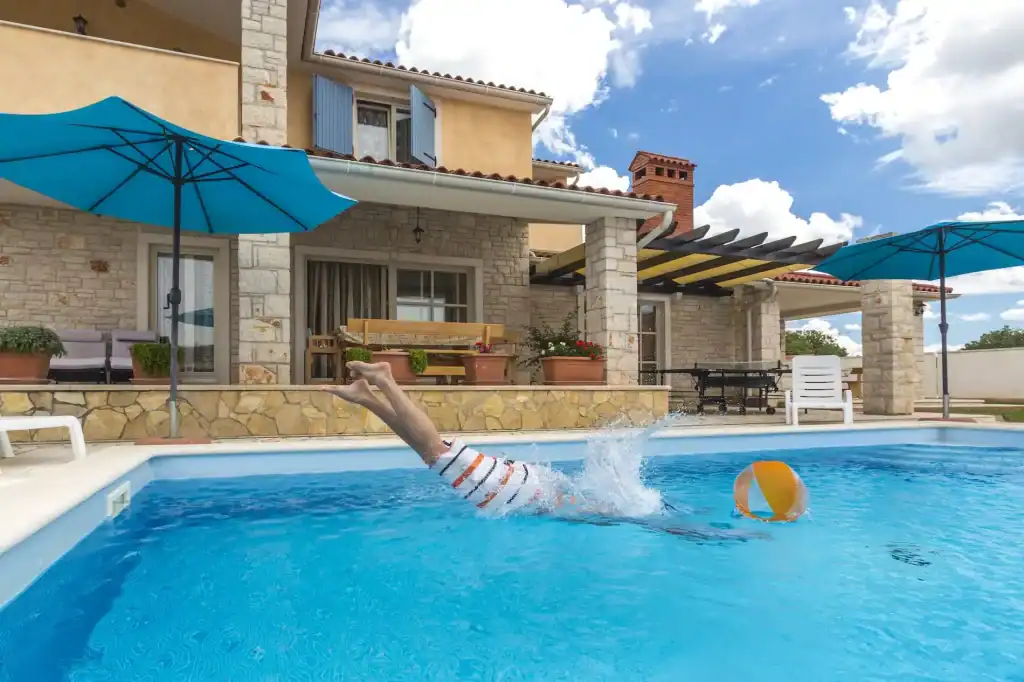 Elegant backyard pool with a child diving in, shaded by blue umbrellas, surrounded by patio furniture, potted plants, and a beautiful stone-trimmed house, perfect for relaxing and entertaining.