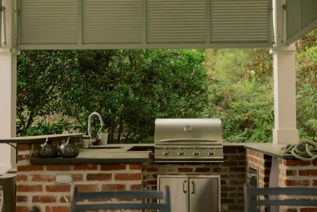 Modern outdoor kitchen with stainless steel grill, sink, and brick countertop under a shaded patio, perfect for poolside entertaining and relaxing.
