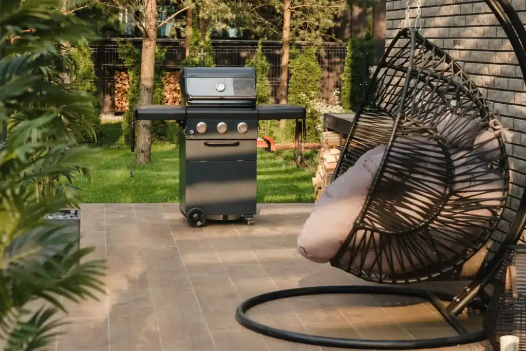 Modern backyard patio featuring a black gas grill, stylish hanging egg chair with cushions, firewood, and lush green trees in the background, perfect for outdoor leisure and BBQs.