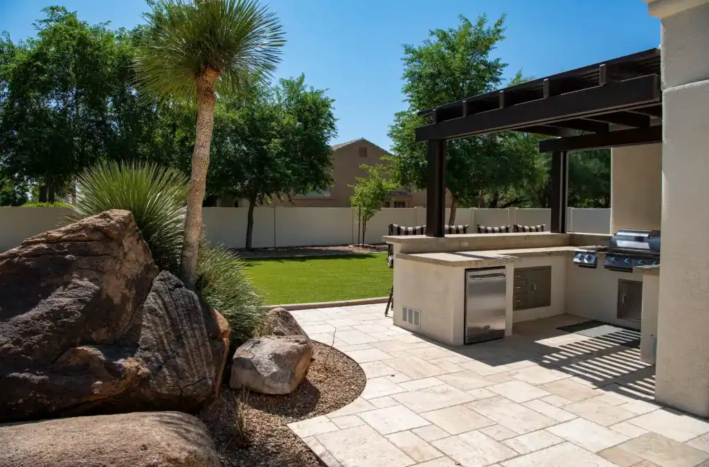 Outdoor kitchen with built-in grill and seating area, surrounded by lush greenery, palm trees, and a well-maintained lawn, ideal for outdoor entertaining and backyard relaxation.