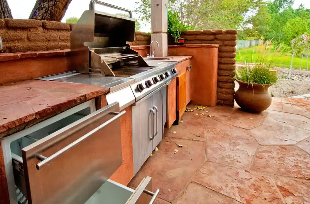 Outdoor kitchen with stainless steel grill, stone countertops, and lush green backyard, perfect for poolside entertaining and relaxation.