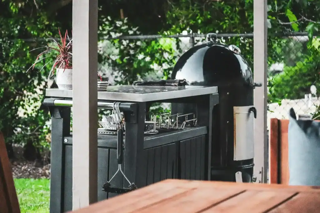Modern outdoor kitchen with a black grill, stainless steel countertop, and green backyard scenery. Perfect for poolside entertaining and enjoying outdoor living spaces.