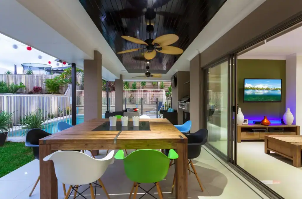Modern outdoor patio with a swimming pool and deck, featuring a wooden dining table, colorful chairs, ceiling fans, and lush landscaping in a backyard setting.