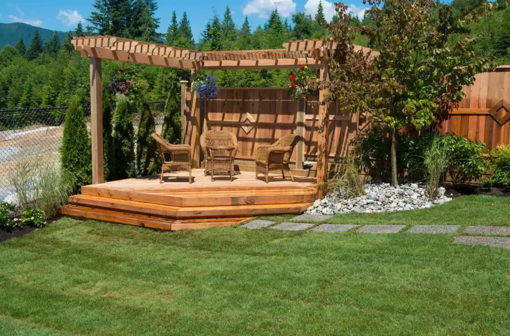 A beautifully crafted wooden deck with a pergola, outdoor seating area, surrounded by lush green lawn, decorative plants, and a scenic backdrop of trees and mountains.