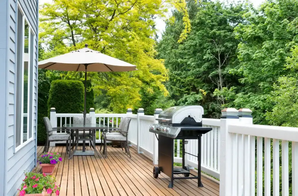 A beautiful wooden outdoor deck with a dining area under a large umbrella, surrounded by lush green trees, featuring a modern barbecue grill and potted flowers, perfect for relaxing summer days.