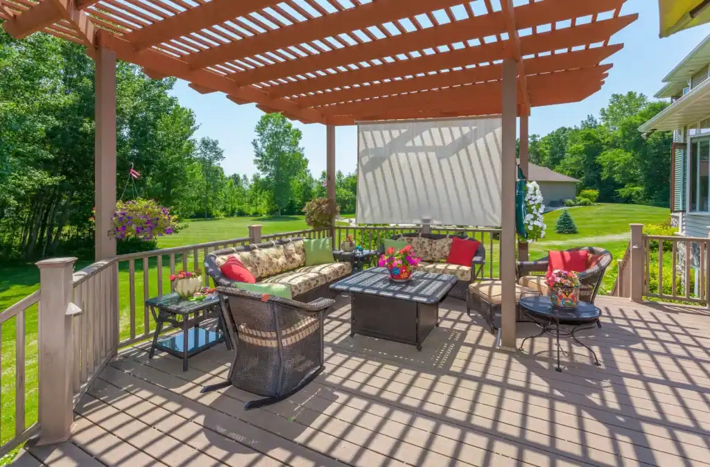 Bright outdoor patio with a pergola, cozy seating area featuring floral cushions and vibrant flower pots, overlooking a lush green backyard with trees and a spacious lawn.