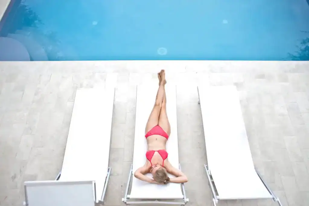 Relaxing woman in a pink bikini lying on a poolside lounge chair by a luxurious blue in-ground swimming pool.
