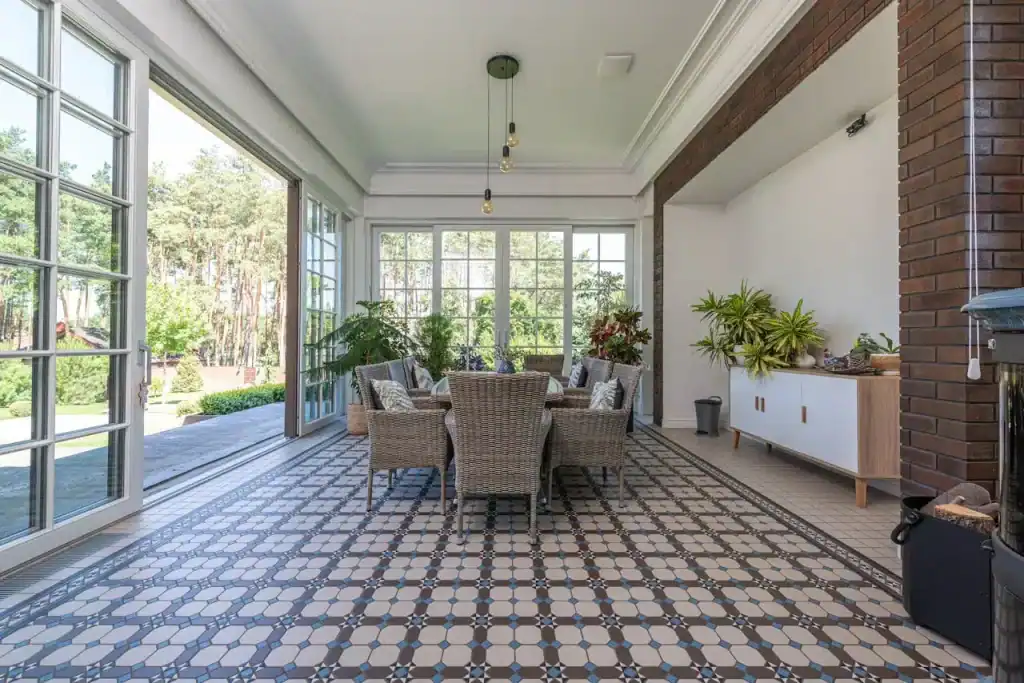 Elegant indoor patio with large glass windows and a view of lush greenery, featuring stylish tiled flooring, wicker dining furniture, and decorative plants for a modern outdoor living space.