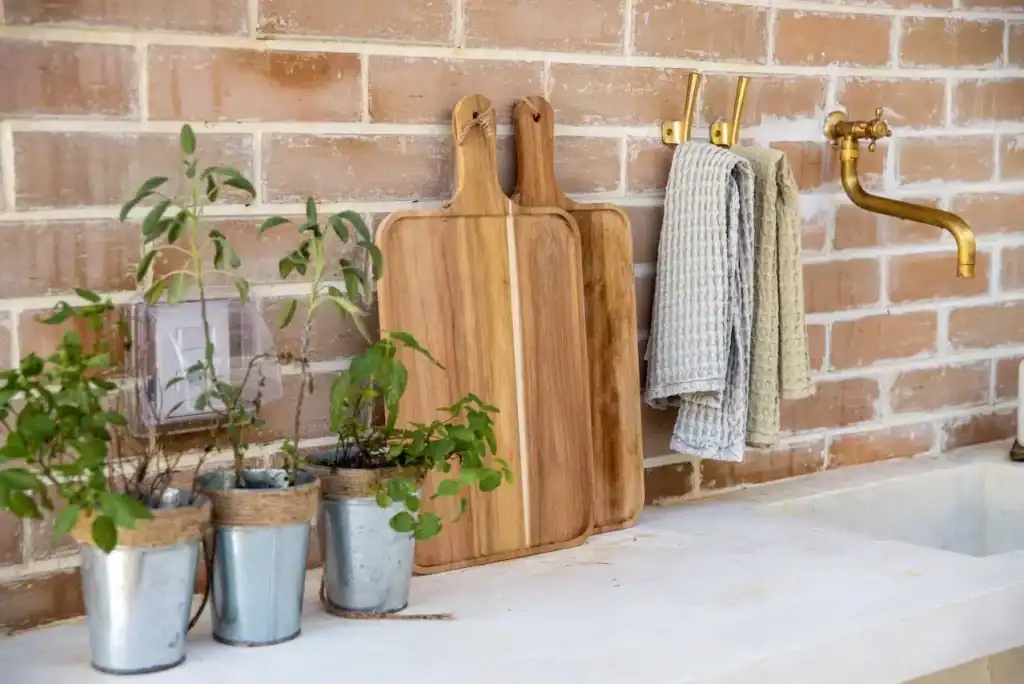 Fresh herbs in metal pots with wooden cutting boards and hanging towels on a brick wall, showcasing a stylish and functional outdoor or indoor laundry or kitchen space.