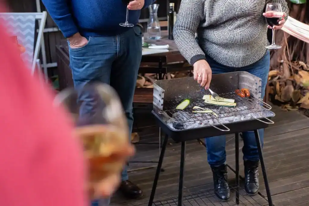 Grilling vegetables & cherries on outdoor BBQ grill during gathering, with people holding wine glasses, on wooden deck, summer evening, casual outdoor party setting.