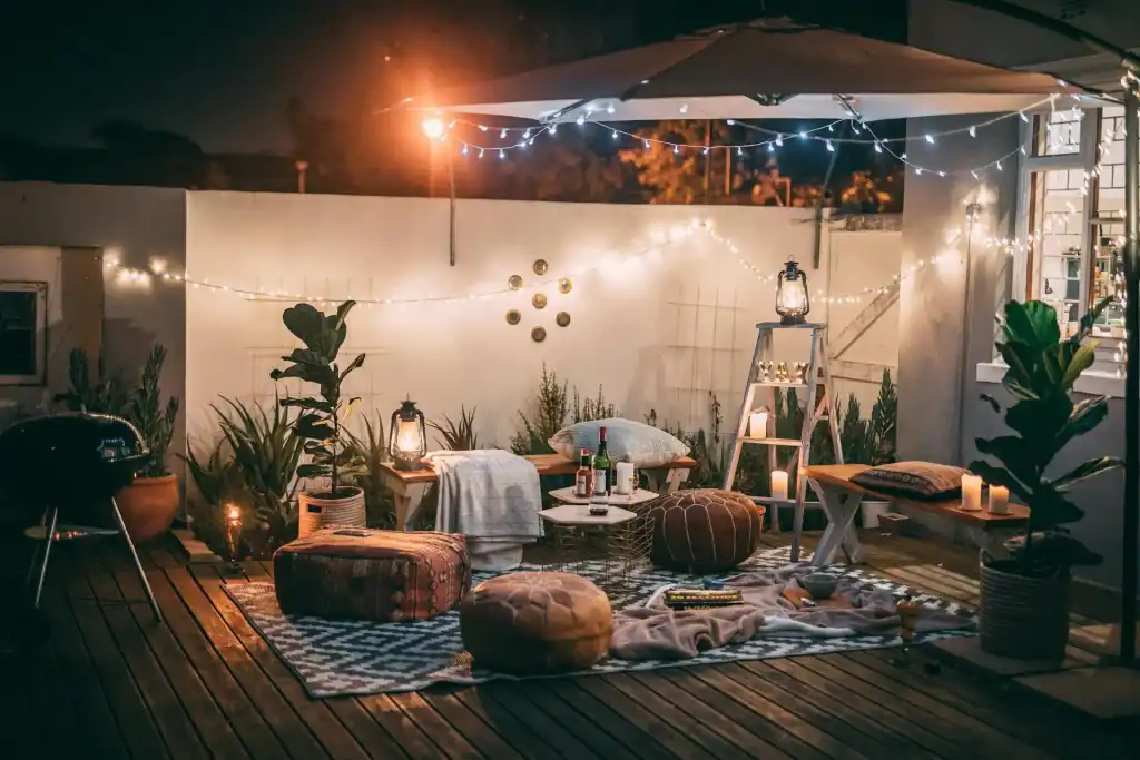 A cozy backyard patio decorated with string lights, lanterns, plush cushions, and plants for an inviting outdoor evening gathering.