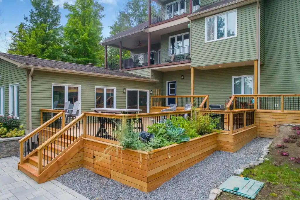 Beautiful multi-level wooden deck with outdoor seating, surrounded by lush greenery and landscaping, connected to a modern green house with large windows and multiple balconies.