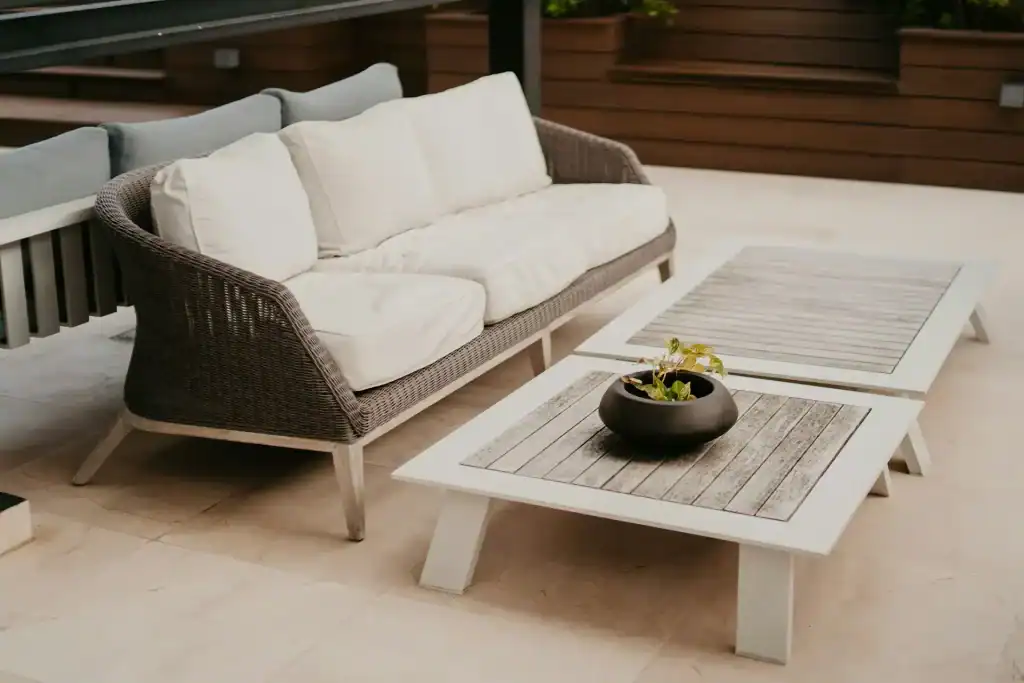 Modern outdoor patio with a cozy wicker sofa with white cushions and two rustic wooden coffee tables, perfect for relaxing and entertaining, featured by Duplessis Builders.