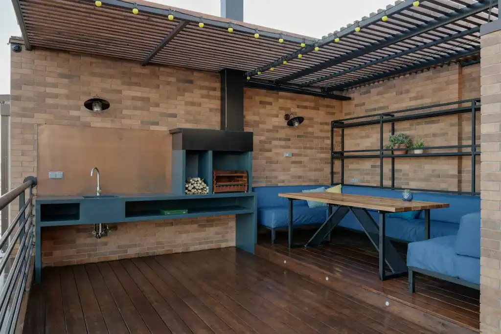 Modern rooftop outdoor kitchen and seating area with brick walls, wooden flooring, built-in grill, and cozy furniture for entertaining.
