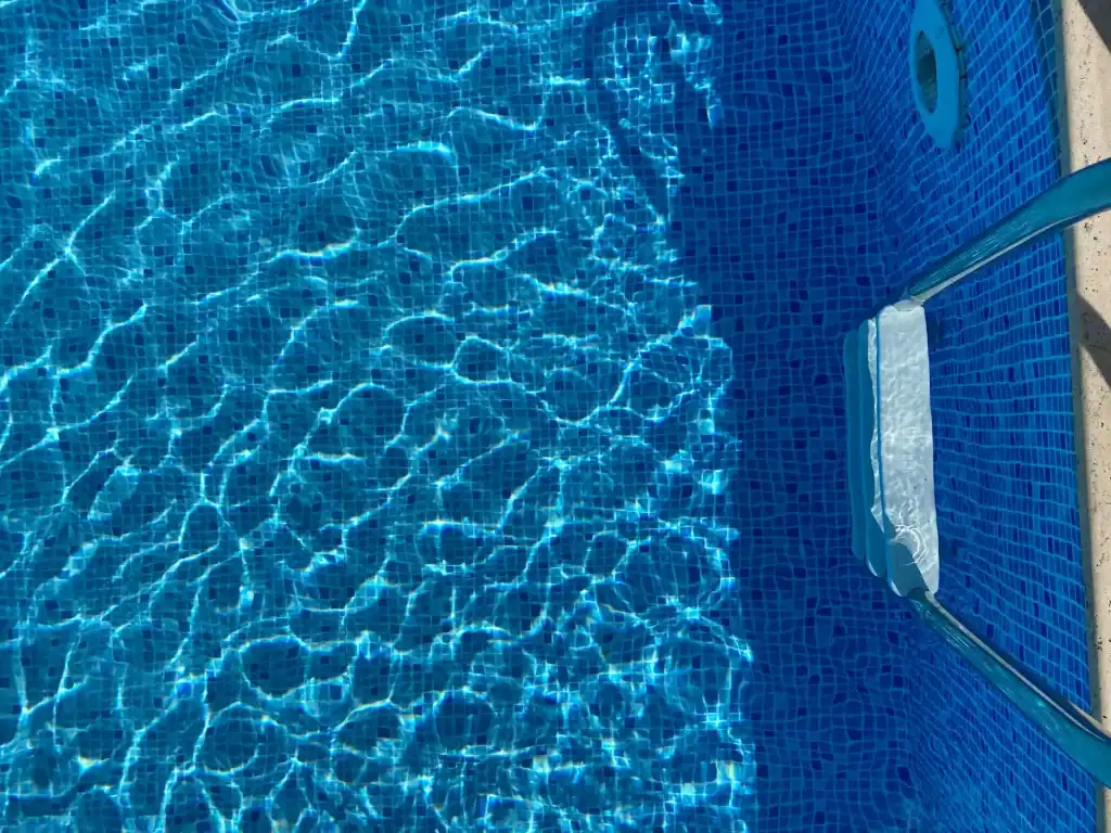 Bright blue mosaic pool tiles with clear, shimmering water reflecting sunlight, featuring a metal ladder for easy access and a pool cover at the edge. Perfectly designed for relaxation and summer fun.