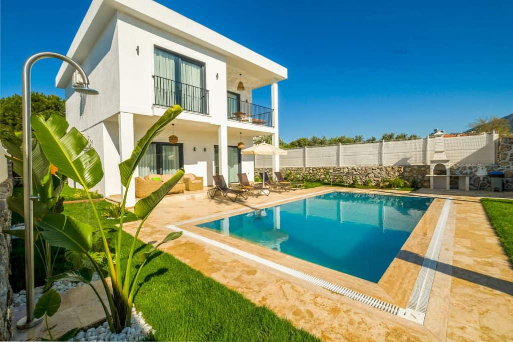 Luxurious modern two-story house with a sparkling backyard pool, outdoor lounging area, and lush greenery on a sunny day. Perfect for relaxing and entertaining in style.