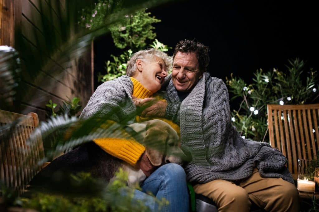 A joyful senior couple laughing and cuddling outdoors at night, enjoying their time together with a dog, surrounded by greenery and warm lighting, emphasizing comfort and happiness after a pool construction project.