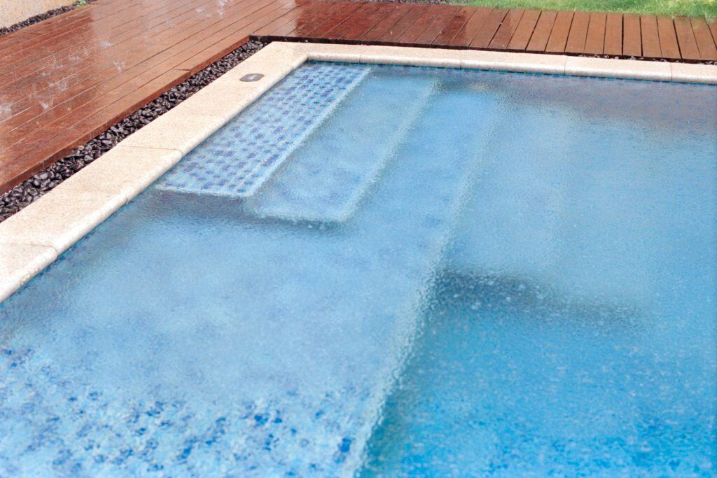A luxury in-ground swimming pool with a built-in hot tub and waterfall, surrounded by a wooden deck, featuring blue water and modern design elements for relaxation and outdoor enjoyment.