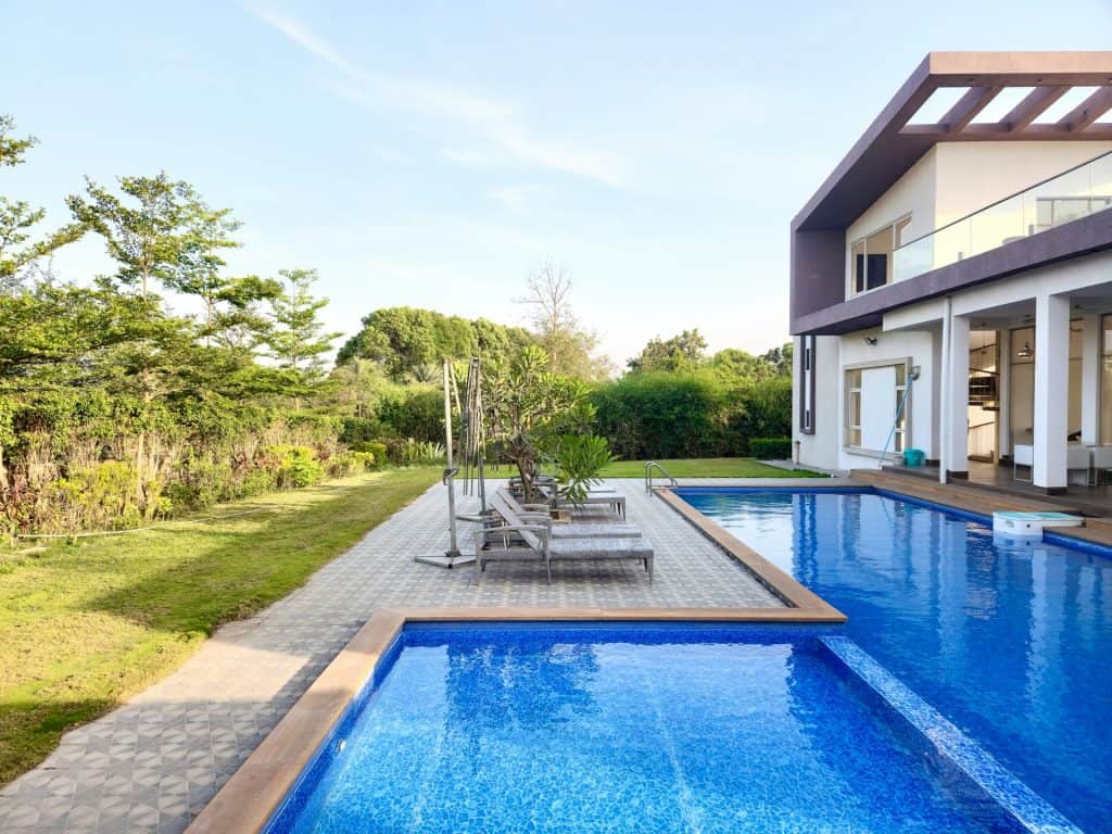 Luxurious modern backyard with a custom in-ground swimming pool, surrounded by lush landscaping and a contemporary two-story house with spacious balconies and outdoor seating areas.