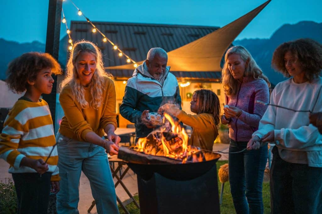 Beautiful backyard gathering around a glowing fire pit with friends and family, under festive string lights, showcasing a custom-designed swimming pool and outdoor living space.