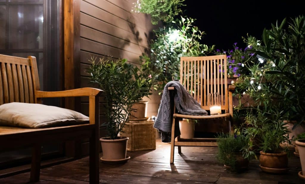 Soft lighting and wooden furniture create a peaceful outdoor relaxing space with plants and cushions.