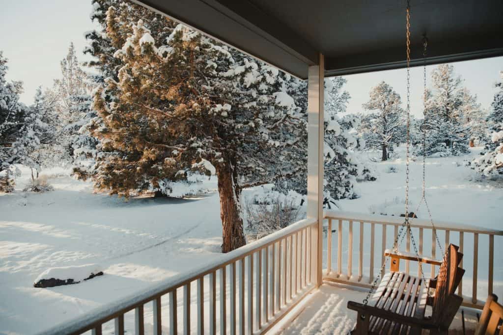 Snow-covered porch with wooden swing overlooking a winter landscape filled with snow-laden pine trees and a peaceful, snowy backyard scene.