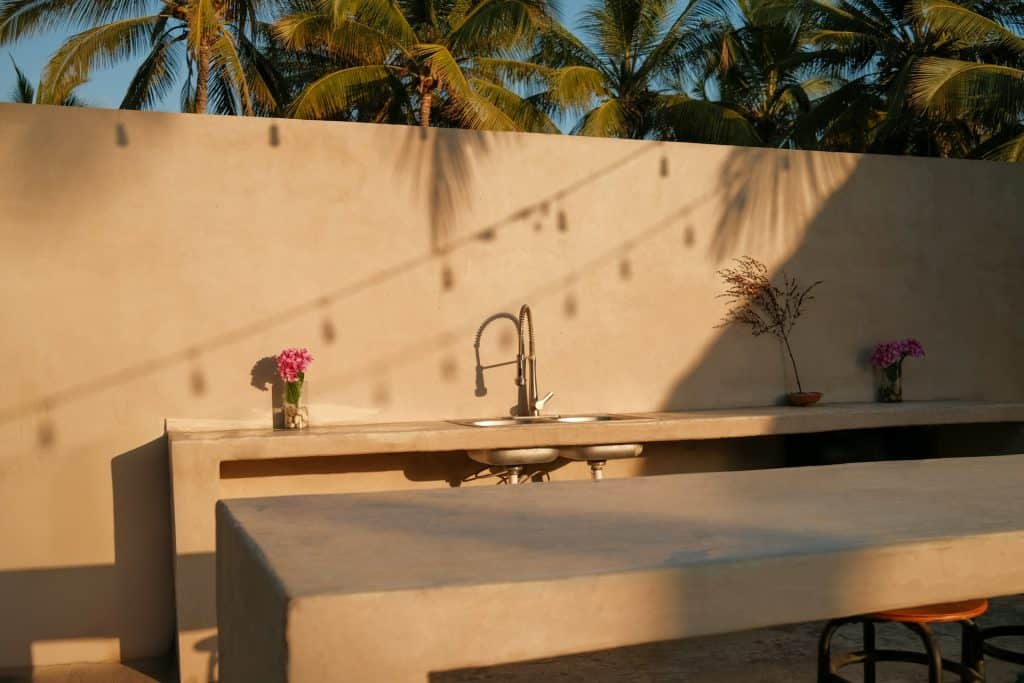 Elegant outdoor poolside kitchen with beige countertops, modern sink, and decorative pink flowers under warm sunlight with tropical palm trees backdrop.