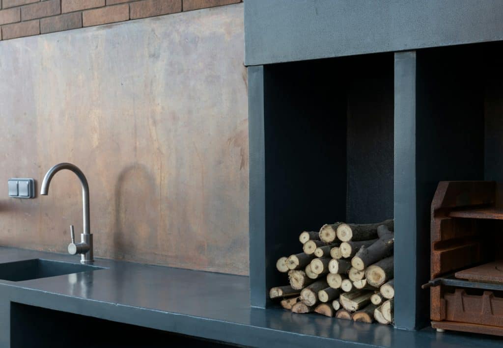 Modern outdoor kitchen featuring black countertop, a stainless steel faucet, and built-in storage for firewood, perfect for stylish outdoor entertainment spaces.