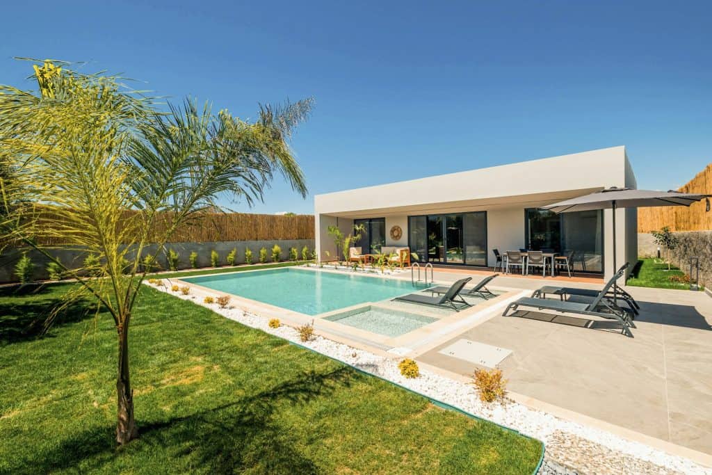 A beautiful backyard featuring a sleek in-ground swimming pool, surrounded by lush green grass, modern patio furniture, and a minimalist architectural house under a clear blue sky.