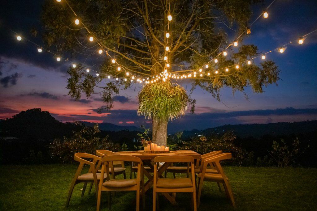 Bright nighttime outdoor scene with string lights hanging from a tree over wooden dining table and chairs, set against a scenic mountain landscape at sunset.