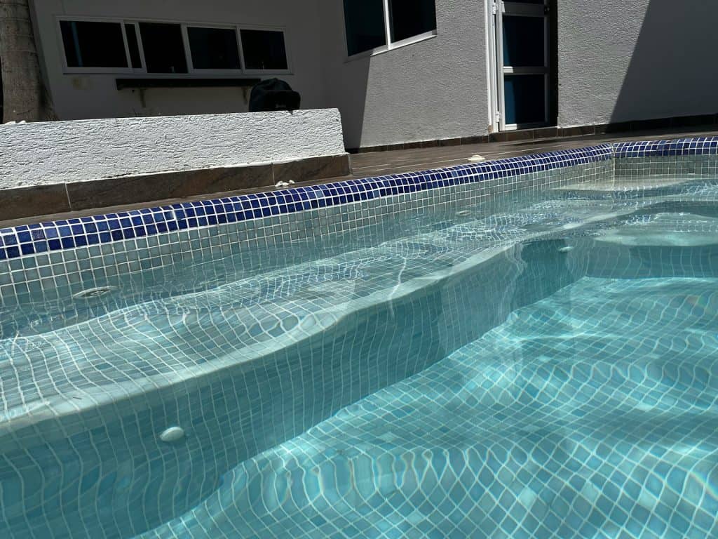A close-up view of a beautifully tiled inground swimming pool with blue mosaic tiles, crystal-clear water, and built-in steps, showcasing high-quality pool installation in a residential backyard.