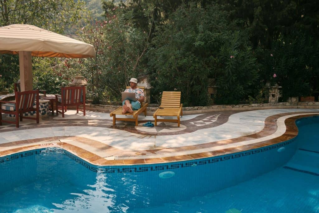 Relaxing outdoor pool scene with lounge chairs, umbrella, lush greenery, and a person working on a laptop, showcasing premium pool installation by Duplessis Builders.