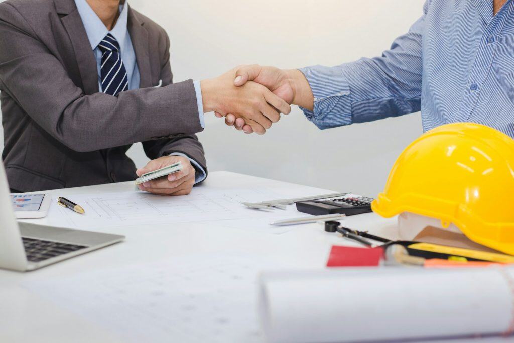 An image of two professionals shaking hands over a contract, with construction plans and a yellow safety helmet on the table, illustrating contract negotiations for pool construction projects.